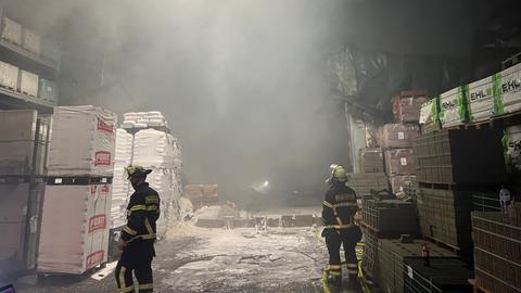 Zwei Feuerwehrleute stehen vor einer Lagerhalle, der Boden ist voller weißer Schaum.