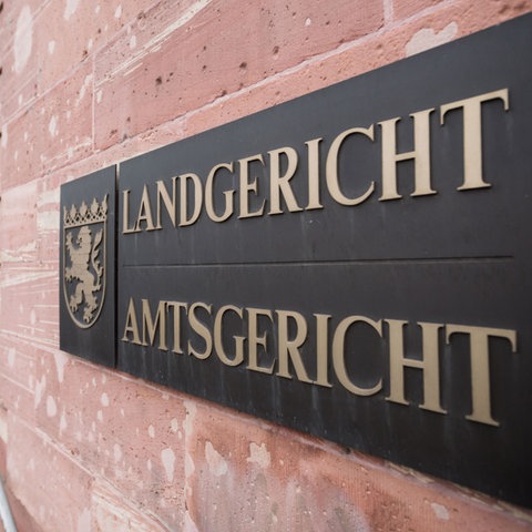 Im Bildvordergrund Schild mit der Aufschrift "Landgericht Amtsgericht" an einer Sandsteinmauer. Im Hintergund unscharf die Fassade und der EIngang des dazugehörigen Gebäudes.