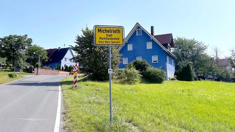 Im Bildvordergrund ein Ortsschild mit der Aufschrift "Michelrieth". Dahinter einzelne kleinere Häuser am Ortseingang.