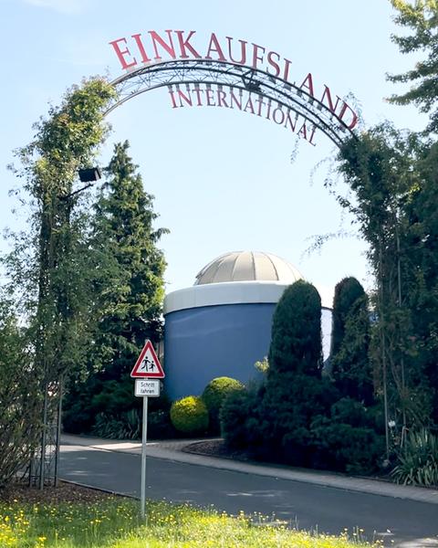 Ein von Kletterpflanzen umranktes Tor mit Schriftzug "Einkaufsland International", dahinter ein kleines blaues Gebäude