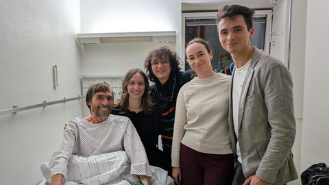 Thomas Weber sitzt im Krankenhaushemd im Bett, daneben seine Familie