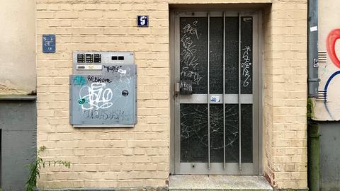 Ein grauer Briefkasten und eine Metalltür mit Glaseinsatz an einer beige gestrichenen Backsteinwand. Beide sind mit Graffiti bedeckt. Oben am Briefkasten sind mehrere Klingelknöpfe mit Namensschildern. Links neben der Tür sind zwei kleine Schilder: eines zeigt "S 47 18", und das andere ein Symbol. Unter der Tür wachsen Unkrautpflanzen aus dem Gehweg. Ein graues Fallrohr verläuft rechts von der Tür an der Wand entlang.