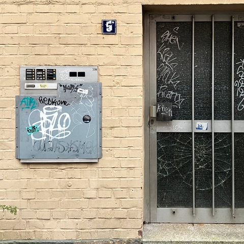 Ein grauer Briefkasten und eine Metalltür mit Glaseinsatz an einer beige gestrichenen Backsteinwand. Beide sind mit Graffiti bedeckt. Oben am Briefkasten sind mehrere Klingelknöpfe mit Namensschildern. Links neben der Tür sind zwei kleine Schilder: eines zeigt "S 47 18", und das andere ein Symbol. Unter der Tür wachsen Unkrautpflanzen aus dem Gehweg. Ein graues Fallrohr verläuft rechts von der Tür an der Wand entlang.