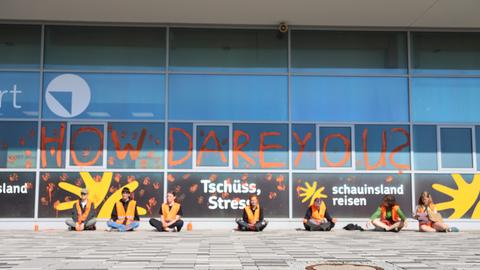 Sechs Mitglieder der Gruppierung "Letzte Generation" sitzen vor dem Flughafen Kassel-Calden vor dem Aufgesprühten Shriftzug "How Dare You?"