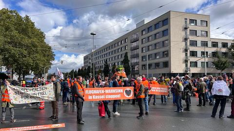 Aktivistinnen und Aktivisten in orangefarbenen Warnwesten stehen auf einer Kreuzung in Kassel und halten Plakate hoch, auf denen steht "Flughafenwahnsinn stoppen"