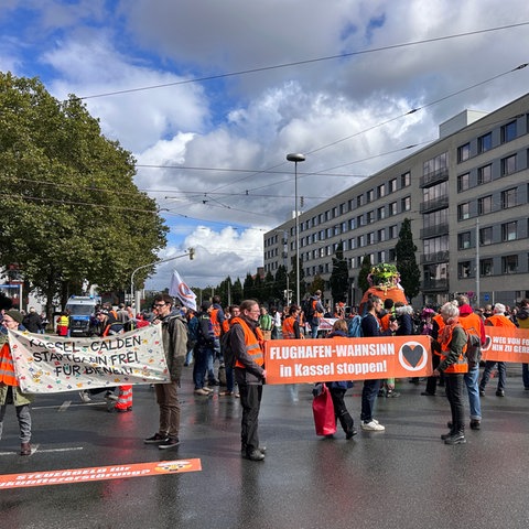 Aktivistinnen und Aktivisten in orangefarbenen Warnwesten stehen auf einer Kreuzung in Kassel und halten Plakate hoch, auf denen steht "Flughafenwahnsinn stoppen"