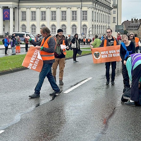 Menschen mit orangfarbenen Bannern - Aufschrift "Weg von Fossil-hin zu gerecht" - stehen verteilt auf einer mehrspurigen Straße mitten auf der Fahrbahn.