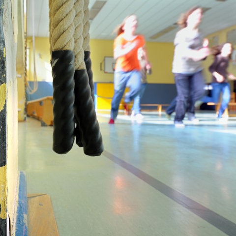 Im Vordergrund verschwindet ein weißes, mit Rostflecken übersähtes Heizungsrohr in einer Wand, von der der Putz abblättert. Im Hintergrund erkennt man, Kinder, die durch eine Turnhalle rennen. 