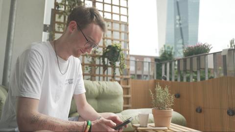 Max Klink sitzt auf seinem Balkon und schaut auf sein Smartphone.