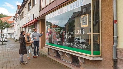 Drei Personen stehen vor dem Laden, auf dem "Leerstandslabor" geschrieben steht. Ein Mann zeigt dabei auf die Scheibe.