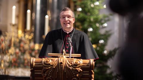 Ein Mann in kirchlicher Amtsrobe steht an einem Pult und spricht. Im Hintergrund unscharf eine Kirche mit Weihnachtsbaum und Kerzen.