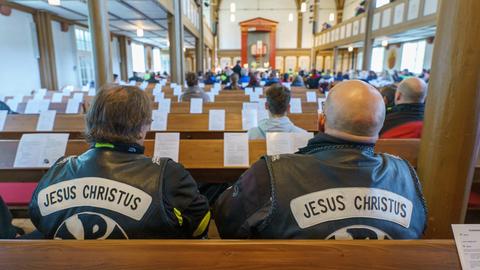 Zwei Motorradfahrer sitzen in einer Kirchenbank - sie tragen Lederkutten mit dem Aufdruck "Jesus Christus".