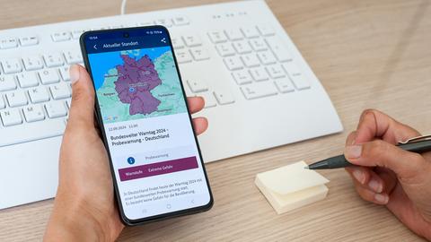 Eine Hand hält ein Smartphone, welches eine eingefärbte Karte von Deutschland mit einem Punkt in Hessen zeigt. Darunter steht "Probewarnung: Warnstufe Extreme Gefahr".