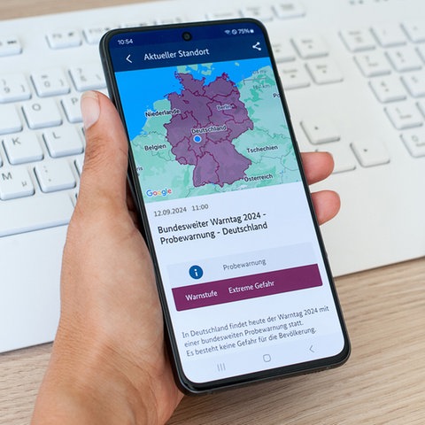 Eine Hand hält ein Smartphone, welches eine eingefärbte Karte von Deutschland mit einem Punkt in Hessen zeigt. Darunter steht "Probewarnung: Warnstufe Extreme Gefahr".