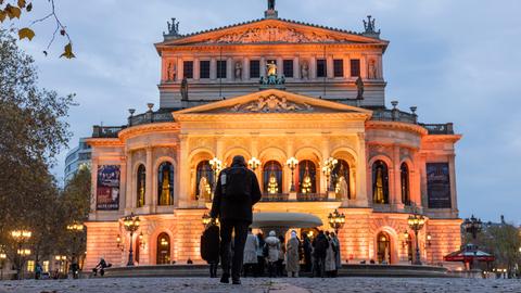 Blick auf Alte Oper in Frankfurt, die orange angestrahlt ist. Im Vordergrund läuft ein Mann mit dem Rücken zur Kamera Richtung Oper.