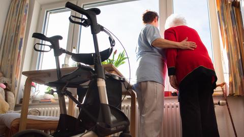 Eine Frau im Kittel und eine ältere Frau schauen zusammen aus einem Fenster im Wohnbereich des Pflegeheims. 