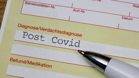 Nahaufnahme Krankenschein, auf welchem unter der Rubrik "Diagnose" "Post Covid" steht. Daneben liegt ein Kugelschreiber.