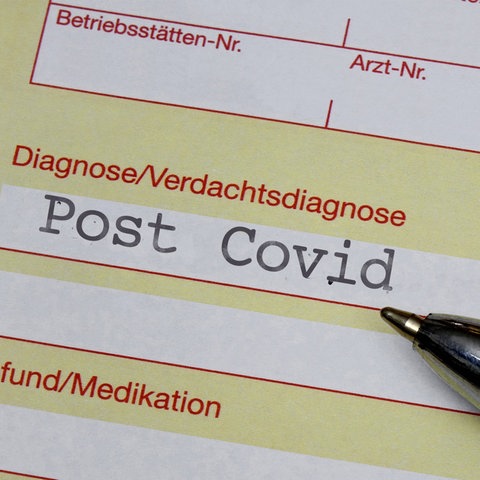 Nahaufnahme Krankenschein, auf welchem unter der Rubrik "Diagnose" "Post Covid" steht. Daneben liegt ein Kugelschreiber.