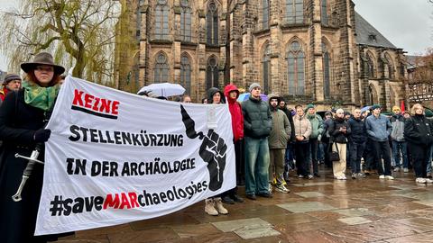 Protestplakat mit Schriftzug: Keine Stellenkürzungen in der Archäologie. Dahinter mehrere Demo-Teilnehmende