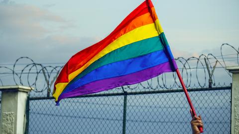 Eine HAnd schwenkt eine Regenbogenfahne vor einem Zaun mit Stacheldraht