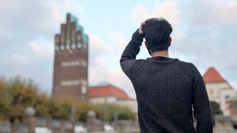 Mensch, der sich an seinen Kopf greift - von hinten fotografiert. Im Hintergrund unscharf ein urbanes Umfeld mit der "Mathildenhöhe".