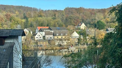 Mehrere Fachwerkhäuser am Ufer eines Flusses, von der anderen Flussseite aus fotografiert, die Bäume tragen Herbstlaub, die Sonne scheint