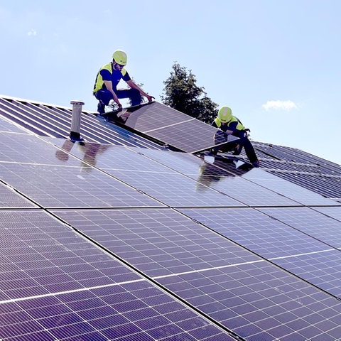 Zwei Männer mit gelben Helmen arbeiten auf einer großen Dachfläche mit vielen Solarpanelen.