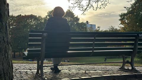 Eine Frau sitzt mit dem Rücken zur Kamera und im Gegenlicht der Sonne auf einer Sitzbank in einem Park