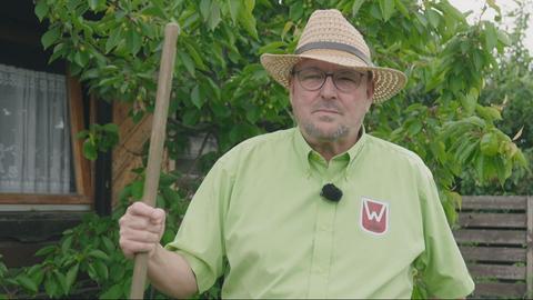 Das Bild zeigt einen älteren Mann in einem hellgrünen Hemd. Er trägt einen Strohhut und hat ein Gartengerät mit langem Holzstiel in der Hand. 