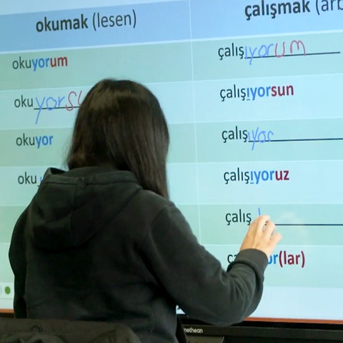 Eine Person (von hinten fotografiert) steht vor einem Smartboard, auf dem Verben der türkischen Sprache in einer Tabelle angeordnet sind. 