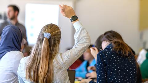 Schülerinnen sitzen mit dem Rücken zur Kamera in einem Klassenzimmer, eine von ihnen zeigt auf. 