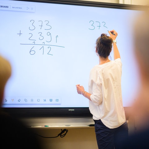 Lehrerin an digitalem Whiteboard macht Matheaufgaben vor Kindern, die sich melden