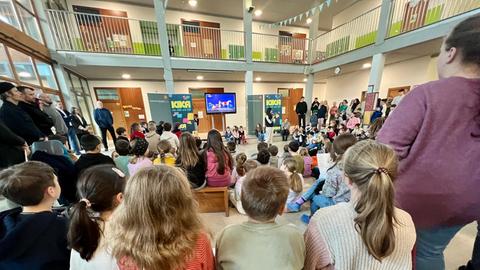 Grundschulkinder von hinten, die in der Schulaula sitzen und in der Mitte einer Frau mit Mikrofron zuhören. Dahinter steht ein großer Fernseher.