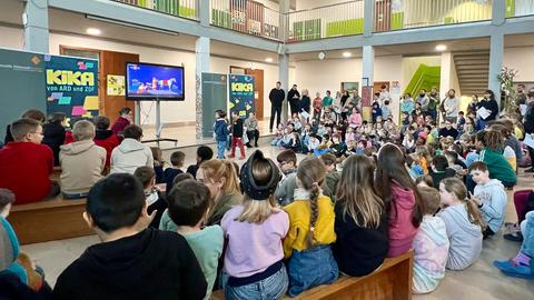 Grundschulkinder von hinten, die in der Schulaula sitzen und in der Mitte einer Frau mit Mikrofron zuhören. Dahinter steht ein großer Fernseher.