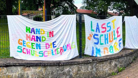 Am Zaun der Schule hängen Transparente gegen die Schließung. Darauf steht in bunter Schrift "Hand in Hand gegen die Schulschließung" und "Lasst die Schule im Dorf".