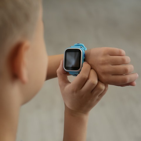 Ein Junge - von hinten fotografiert - guckt auf seine blaue Smartwatch und drückt daran auf einen Knopf