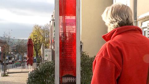 Ein Mann in roter Jacke (von hinten) steht vor einer Betonstele, die einen roten Bereich in ihrer Mitte hat, auf welchem ein langer Text steht. Im Hintergrund eine Geschäftsstraße.