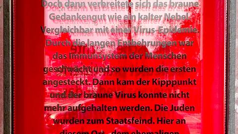 Nahaufnahme: Roter Bereich (Glas mit Spiegelungen), auf welchem ein langer Text in schwarzen Buchstaben steht: "Vergleichbar mit einer Virus-Epidemie. Durch die langen Entbehrungen war das Immunsystem der Menschen geschwächt und so wurden die ersten angesteckt. Dann kam der Kipppunkt und der braune Virus konnte nicht mehr aufgehalten werden. Die Juden wurden zum Staatsfeind. Hier an diesem Ort - dem ehemaligen Adolf-Hitler-Platz - wurde die Familie Höxter ausgegrenzt, beleidigt, angefeindet, geschlagen, gequält, enteignet udn Familienmitglieder deportiert und ermordet. Hier an diesem Platz wurde ihr Zuhause - das Kaufhaus Höxter - im Jahr 1938 enteignet und abgerissen. Der braune Mopp war zufrieden und die Nachbarn."