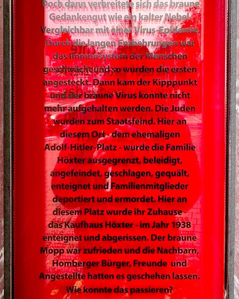 Nahaufnahme: Roter Bereich (Glas mit Spiegelungen), auf welchem ein langer Text in schwarzen Buchstaben steht: "Vergleichbar mit einer Virus-Epidemie. Durch die langen Entbehrungen war das Immunsystem der Menschen geschwächt und so wurden die ersten angesteckt. Dann kam der Kipppunkt und der braune Virus konnte nicht mehr aufgehalten werden. Die Juden wurden zum Staatsfeind. Hier an diesem Ort - dem ehemaligen Adolf-Hitler-Platz - wurde die Familie Höxter ausgegrenzt, beleidigt, angefeindet, geschlagen, gequält, enteignet udn Familienmitglieder deportiert und ermordet. Hier an diesem Platz wurde ihr Zuhause - das Kaufhaus Höxter - im Jahr 1938 enteignet und abgerissen. Der braune Mopp war zufrieden und die Nachbarn."