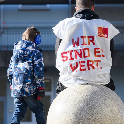 Rückansicht eines Mannes, der auf einem kugelförmigen Stein sitzt und eine weiße Plastikweste trägt, auf welcher in roten Buchstaben "Wir sind es wert" steht. Daneben sitzen zwei Kinder.