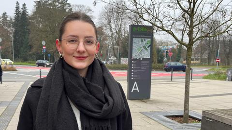 Eine Frau steht auf einem Platz und lächelt in die Kamera. Hinter ihr fahren Autos. Sie hat braune Haare, trägt eine Brille und Ohrringe. Die Haare sind zu einem Zopf gebunden. Sie trägt eine schwarze Jacke und einen schwarzen Schal.