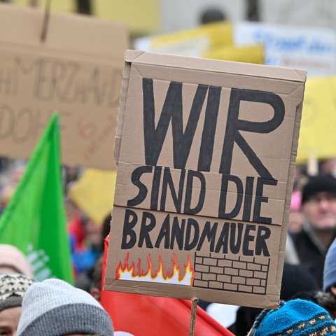 Menschen protestieren gegen rechts, ein Teilnehmer hält ein Pappschild mit der Aufschrift: "Wir sind die Brandmauer".