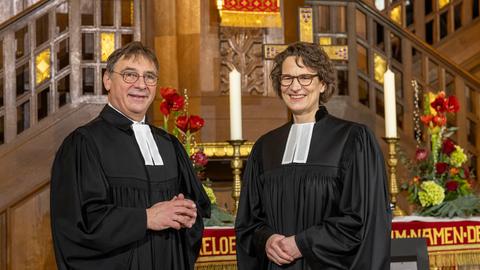 Christiane Tietz (r.) und Volker Jung stehen im Talar vor dem mit zwei Kerzen und Blumengestecken geschmückten Altar in der Wiesbadener Lutherkirche