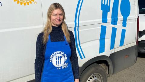 Eine blonde Frau steht vor einem Kleintransporter. Sie trägt eine Schürze mit der Aufschrift "Tafel Frankfurt".