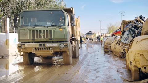 Eine Straße in den spanischen Katastrophengebieten. Am rechten Fahrbahnrand stapeln sich zerstörte Autos im Schlamm. Links fährt ein Militärlaster.