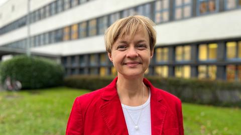 Frau mit kurzen blonden Haaren und rotem Blazer schaut in die Kamera, im Hintergrund sind grüne Büsche und ein weißes Gebäude zu erkennen.