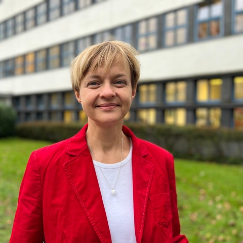 Frau mit kurzen blonden Haaren und rotem Blazer schaut in die Kamera, im Hintergrund sind grüne Büsche und ein weißes Gebäude zu erkennen.