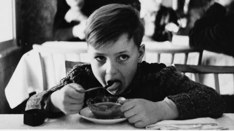 Das Schwarz-weiß-Foto zeigt einen achtjährigen Jungen, der am Tisch sitzt und mit einem Löffel aus einem Teller ist. Im Hintergrund sind unscharf zwei weitere Jungen zu sehen.