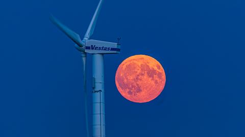 Ein orangfarbener Mond im blauen Nachthimmel, direkt neben dem Mond der obere Teil eines Windrads.