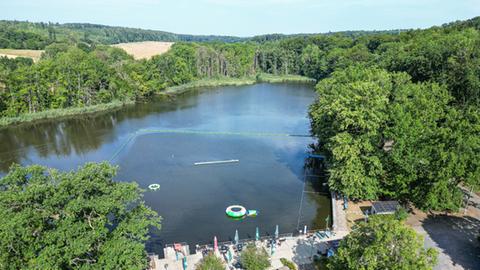 Blick von oben auf ein rechteckiges kleines Schwimmbecken im Wald mit größerem Stausee dahinter